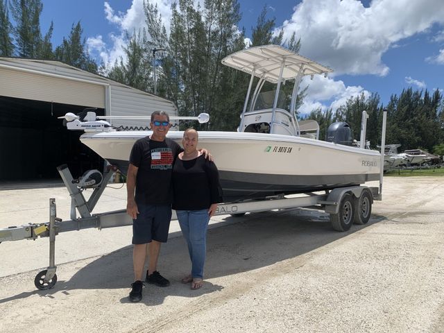 Couple standing beside a white center-console boat on a trailer in a gravel lot under a blue sky