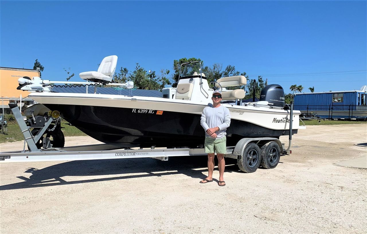Person standing beside a white boat on a trailer in a sunny outdoor lot