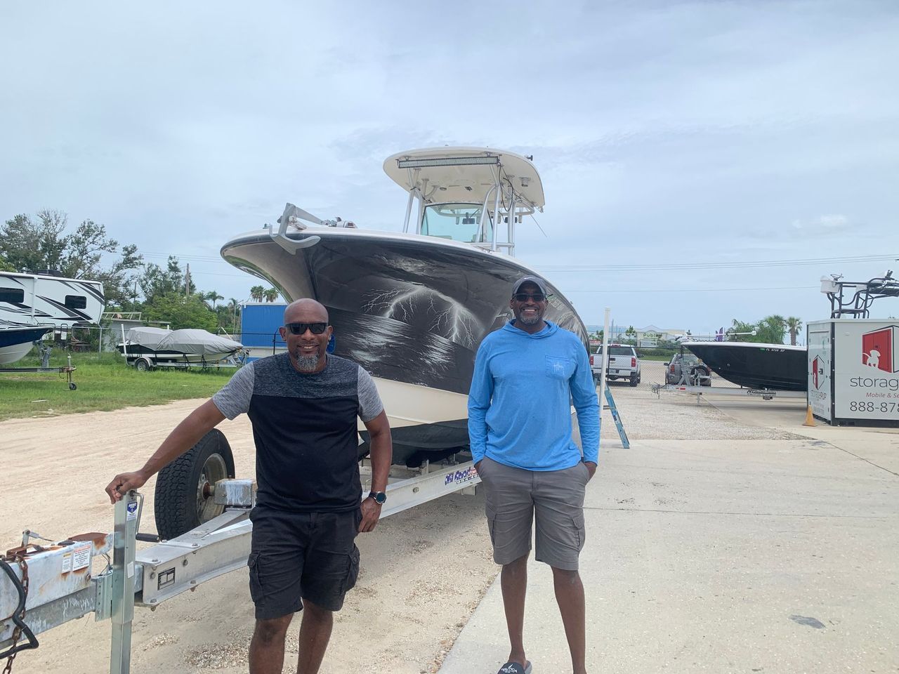 Two people standing beside a large boat at a marina, with a dock and parked vehicles in the background.