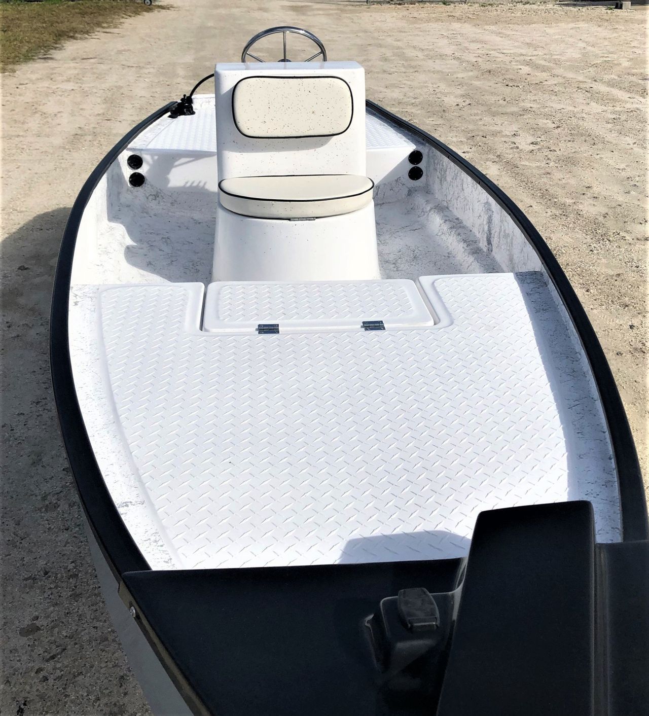 A white center-console boat sits on a gravel surface, showing a textured deck, a single seat, and a steering wheel.