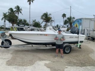 White boat on a trailer with a person standing beside it in a marina lot, palm trees in the background
