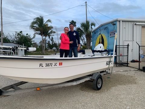 Two people standing on a boat trailer beside a small boat and a building mural under cloudy skies