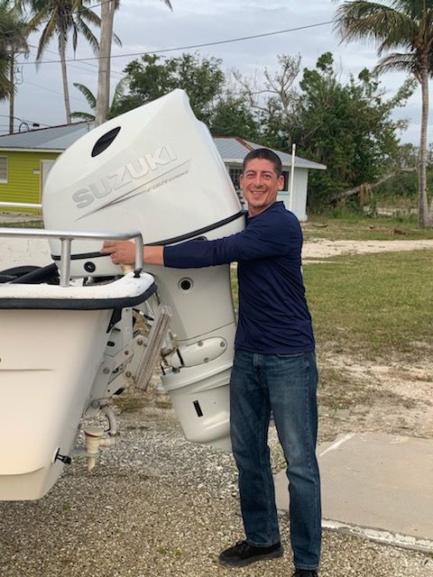 Man smiling while holding a white boat motor beside a boat on a gravel lot