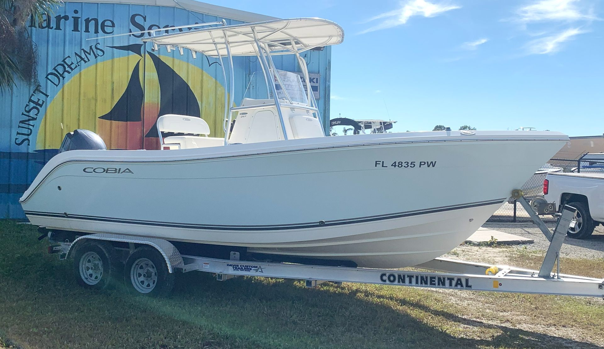 White fishing boat on a trailer outdoors under a blue sky, with a marina sign in the background