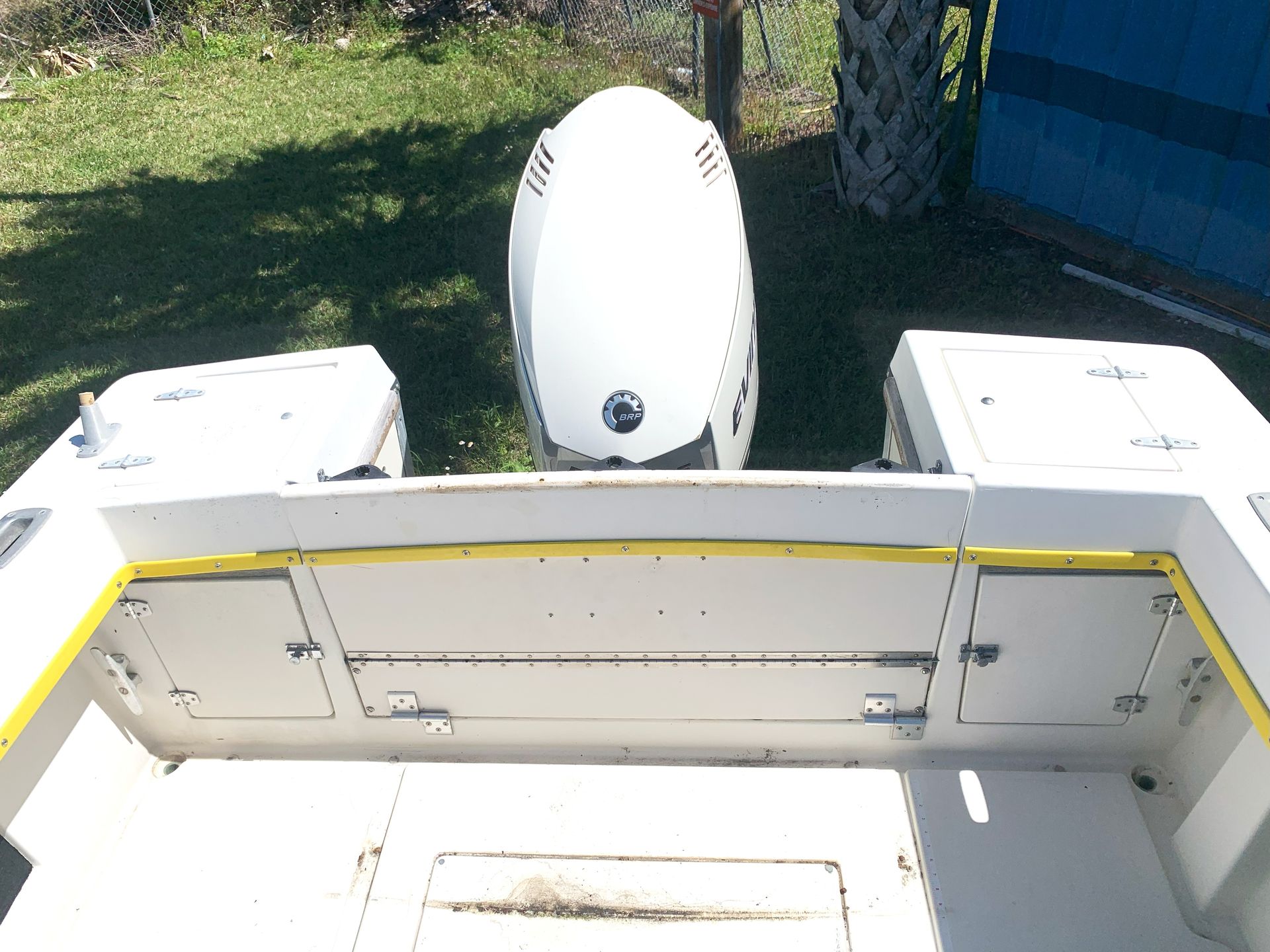 White boat stern with open storage hatch and a small white motor in the background on grass near water