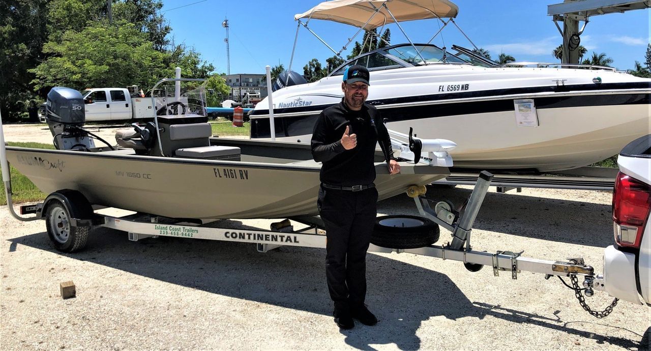 Man standing beside a small tan boat on a trailer in a sunny parking lot, with a larger white boat behind him.