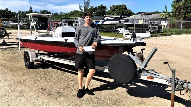 Person standing beside a red-and-black boat on a trailer in a sunny outdoor lot