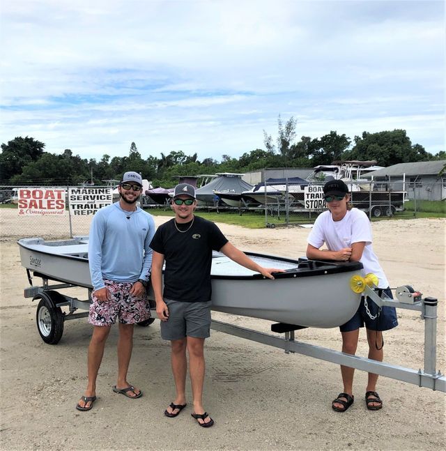 Three people posing beside a small boat on a trailer at an outdoor boat sale lot.