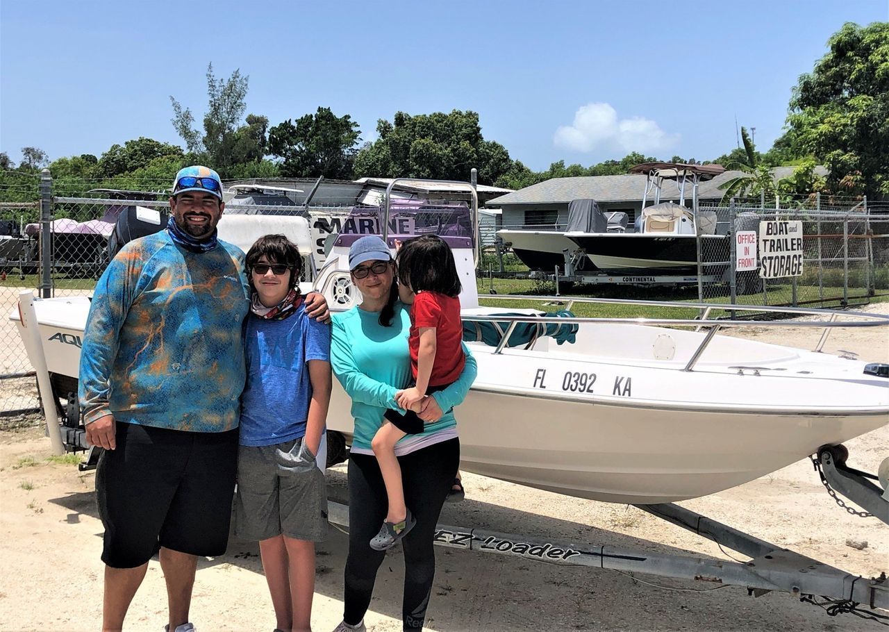 Four people posing by a white boat in a sunny marina, with trees and docked boats in the background
