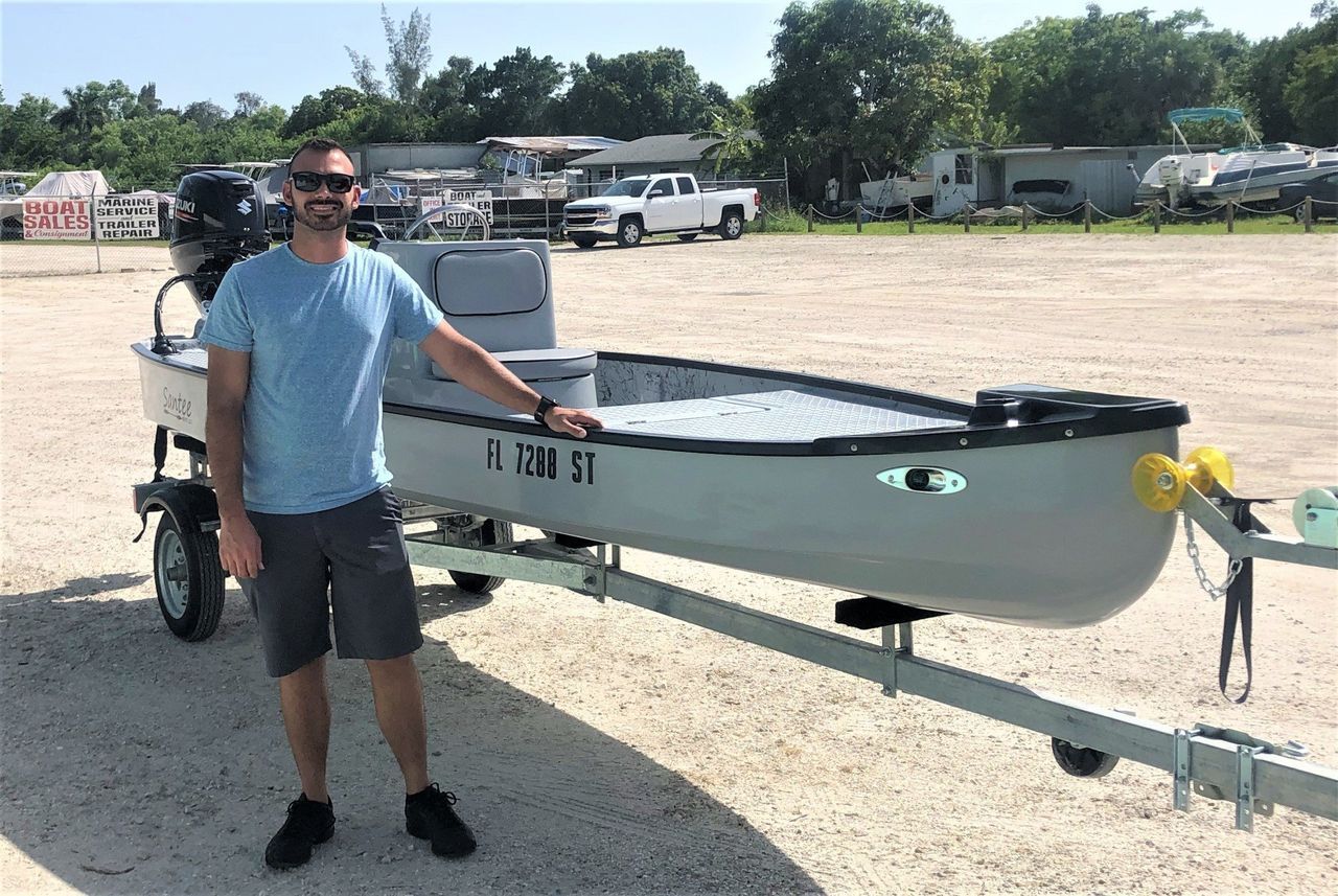 Man standing beside a small white boat on a trailer in a gravel lot under sunny skies