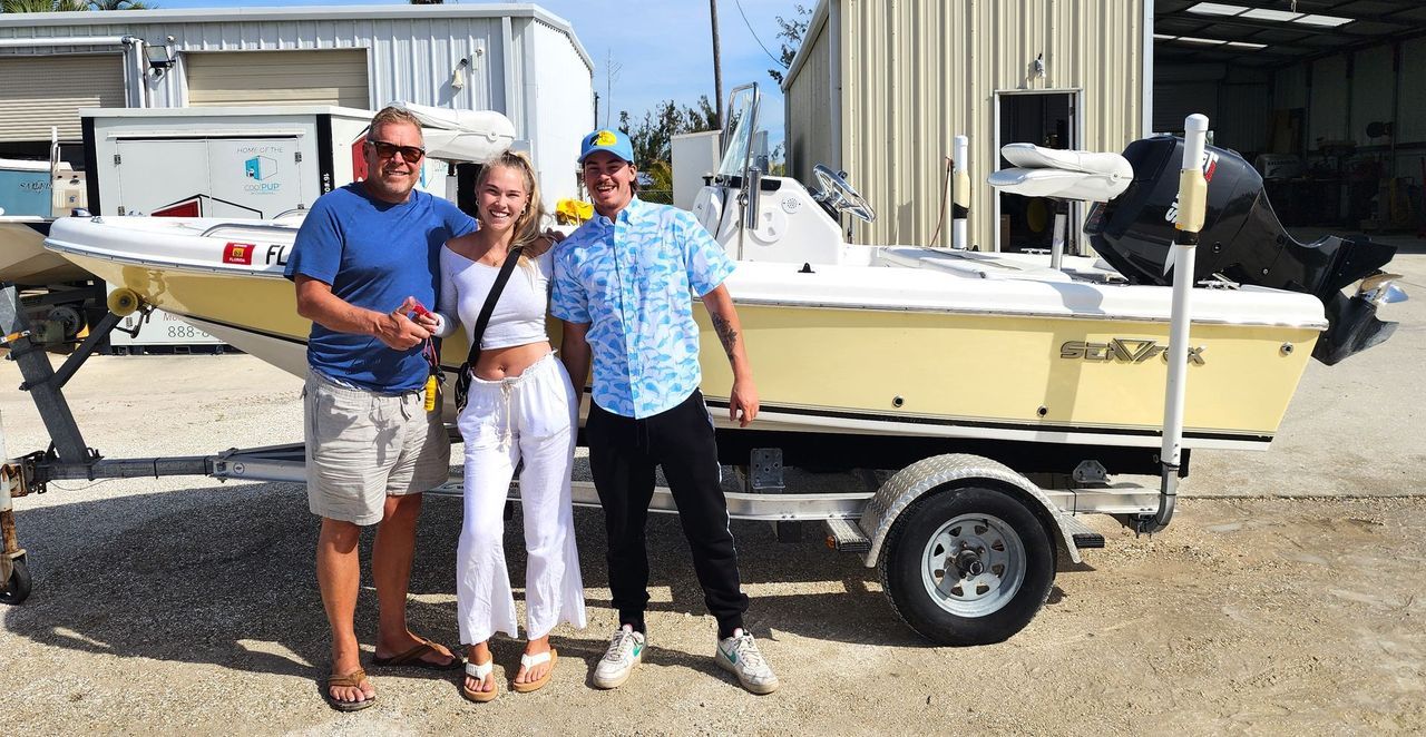 Three people posing in front of a small boat on a trailer in a marina yard.