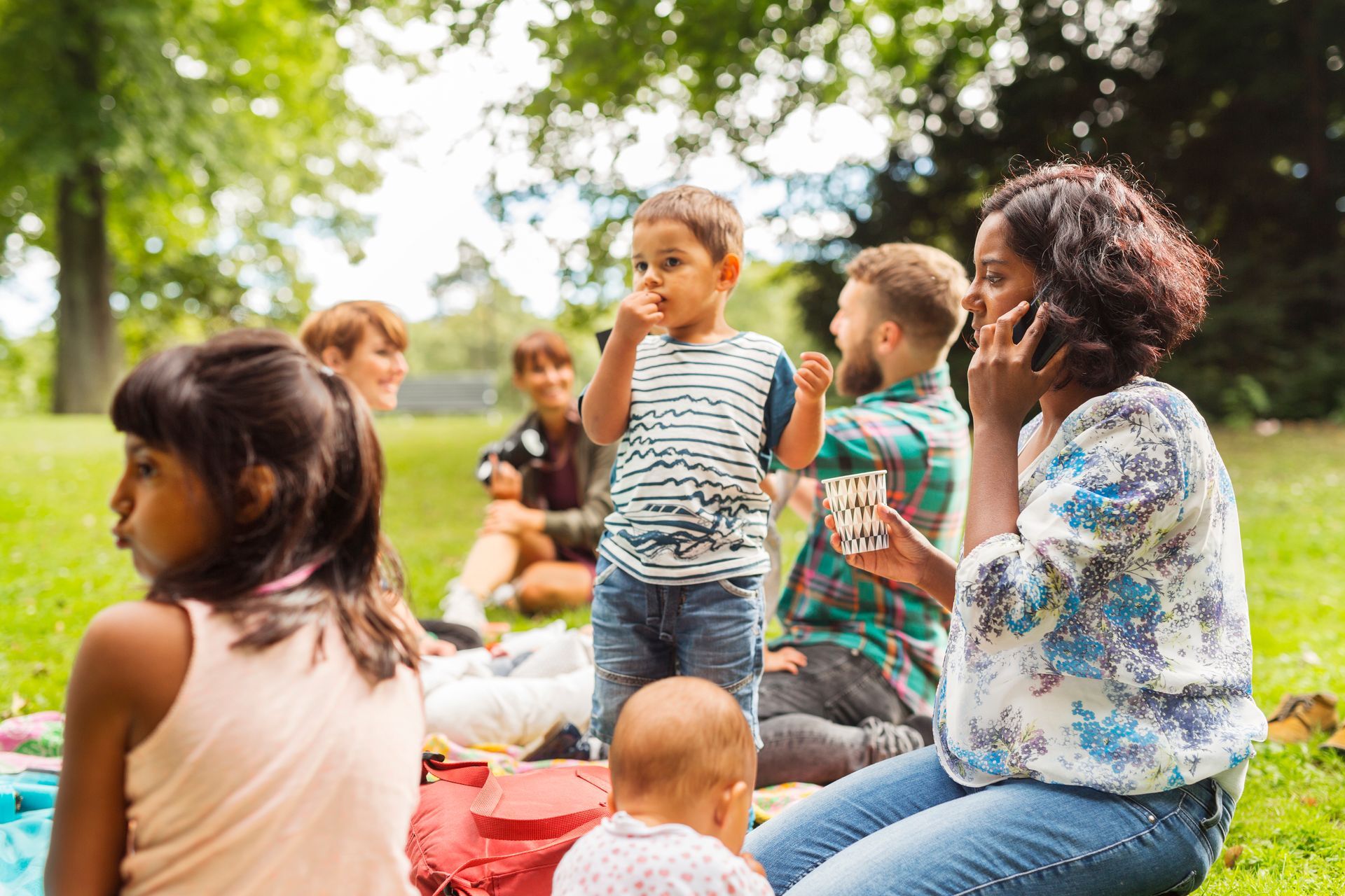Children sit and stand on blankets in a sunny park during an outdoor gathering