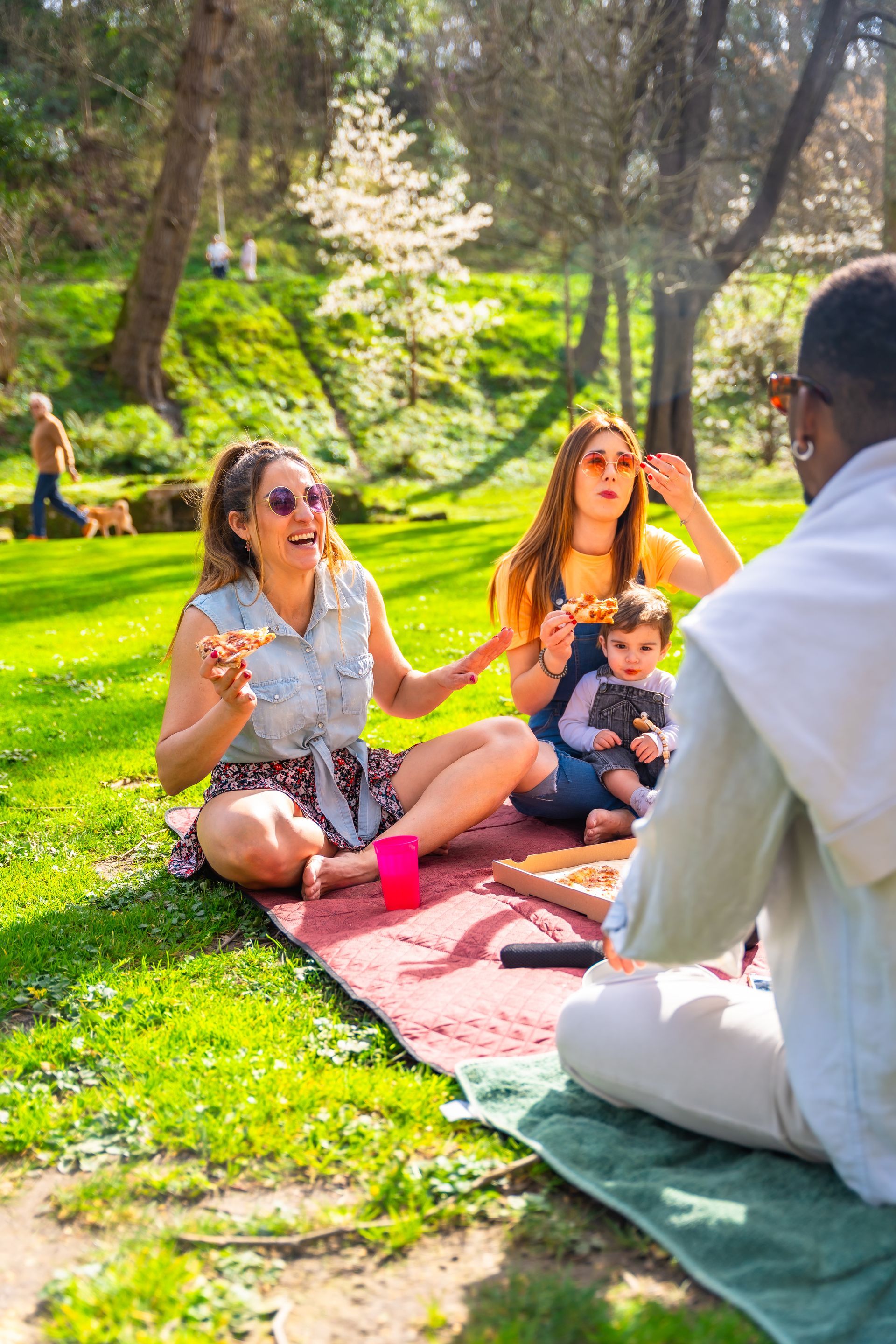 Friends sitting on blankets in a sunny park, smiling and chatting with a baby in the center.
