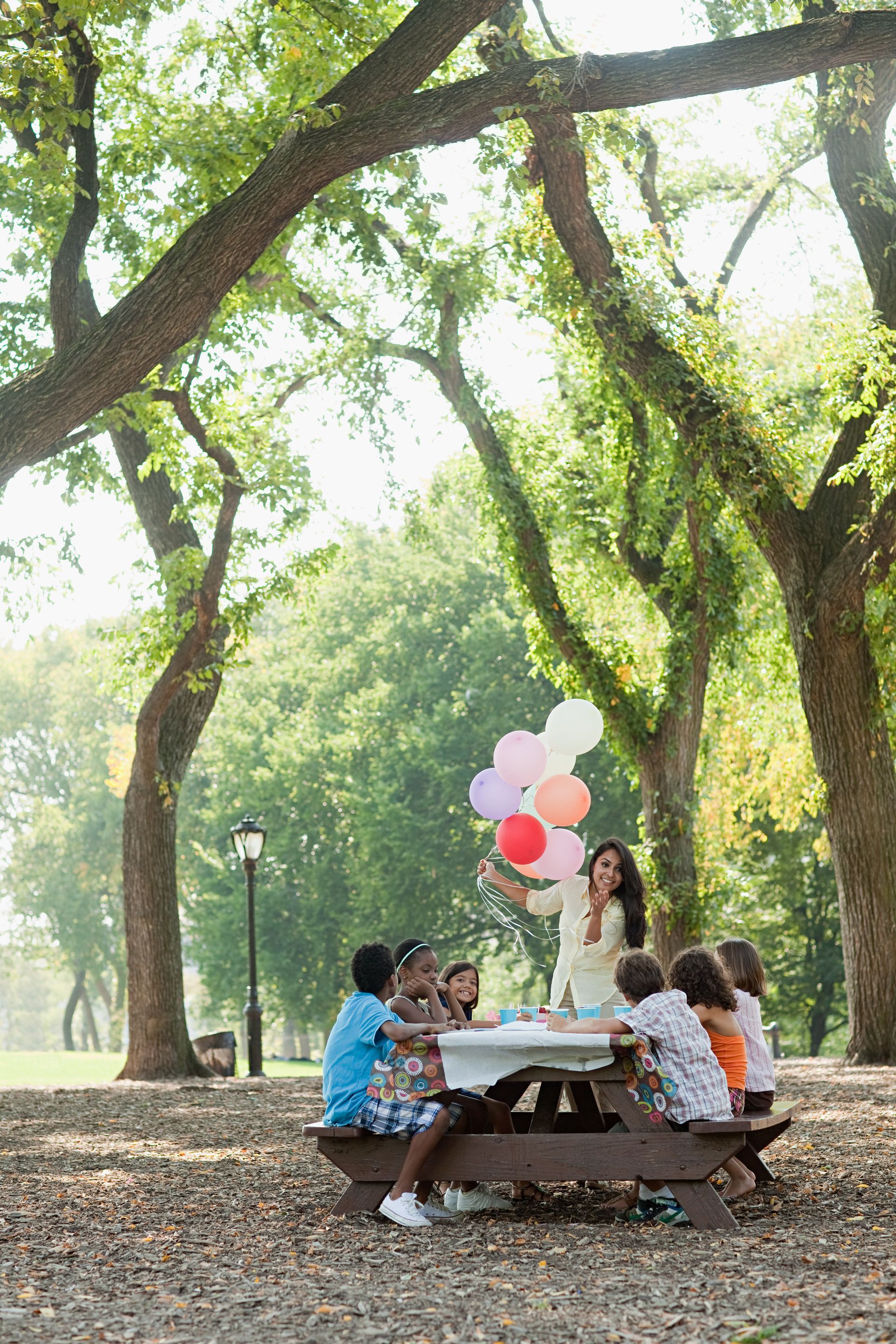 People seated at a picnic table in a sunny park with colorful balloons under large trees