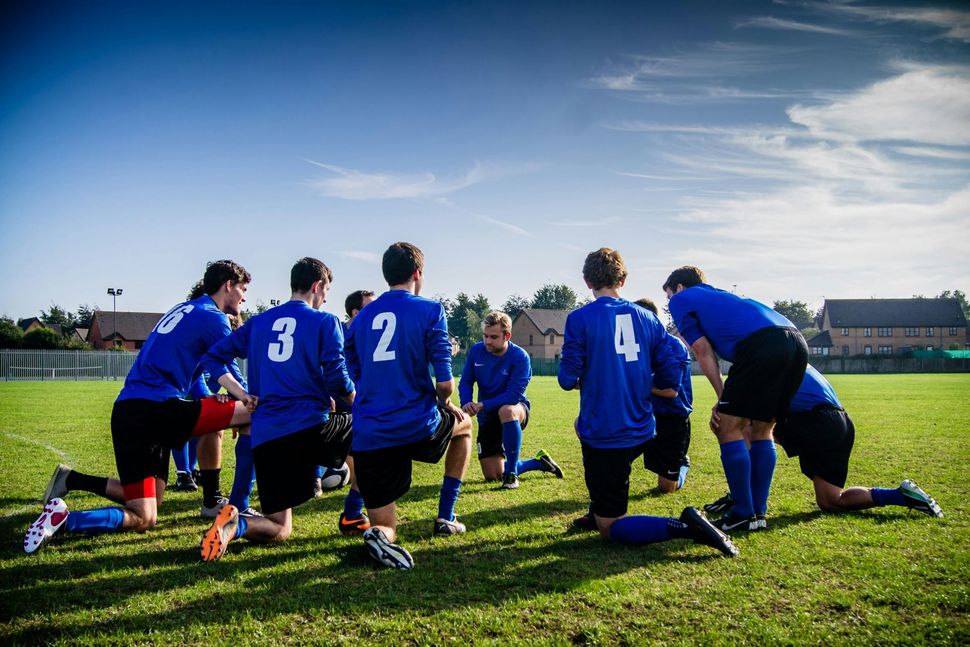 Soccer players in blue jerseys kneel in a huddle on a grassy field before a match