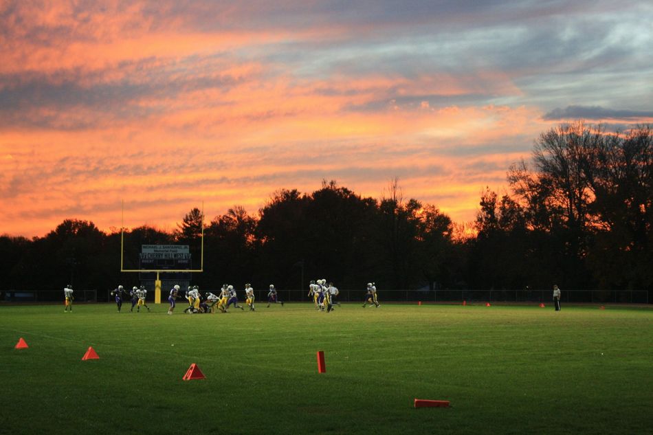 Football players on a field at sunset, with orange cones and silhouetted trees in the background