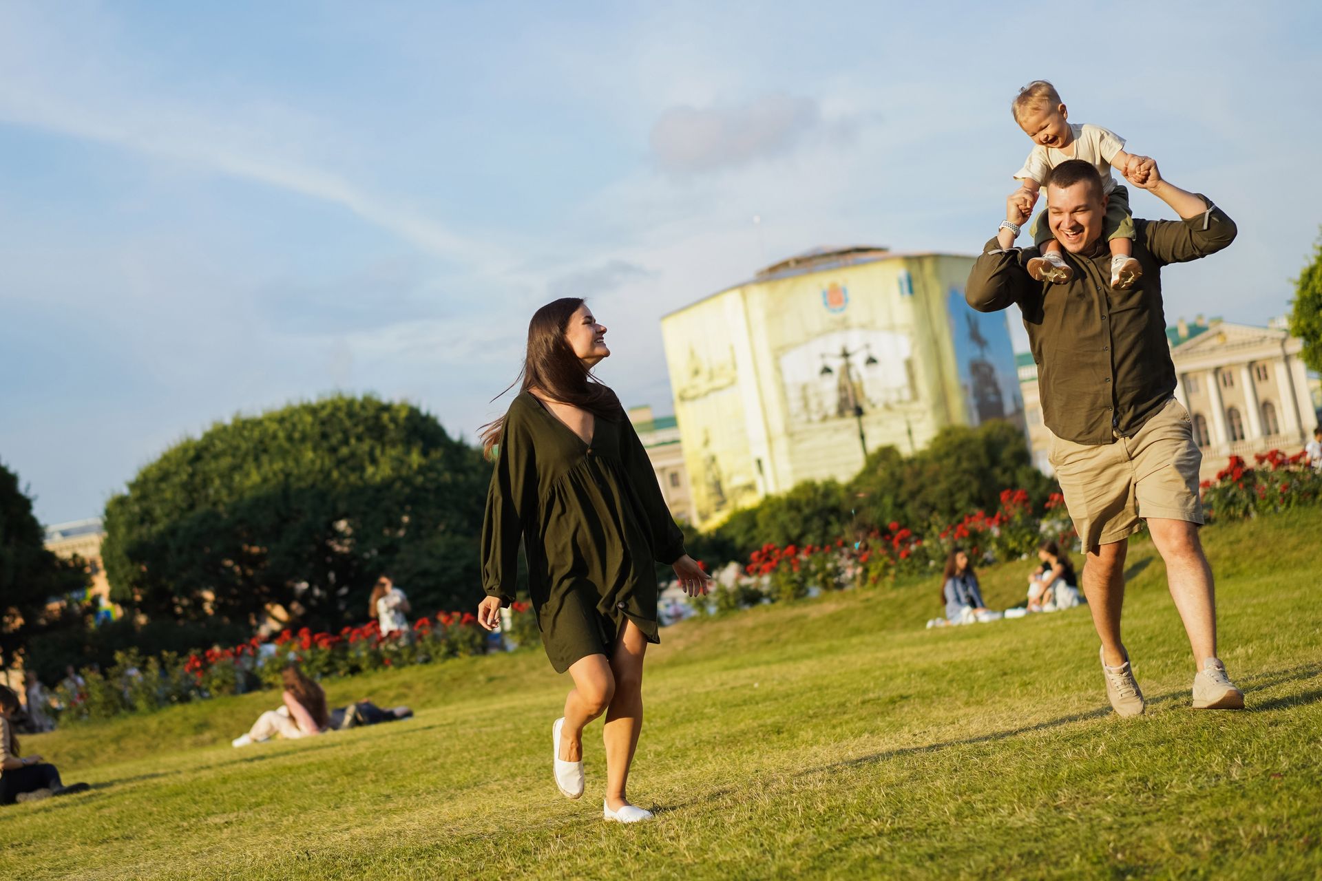 Two people playing in a grassy park, one giving the other a piggyback ride near a large building.