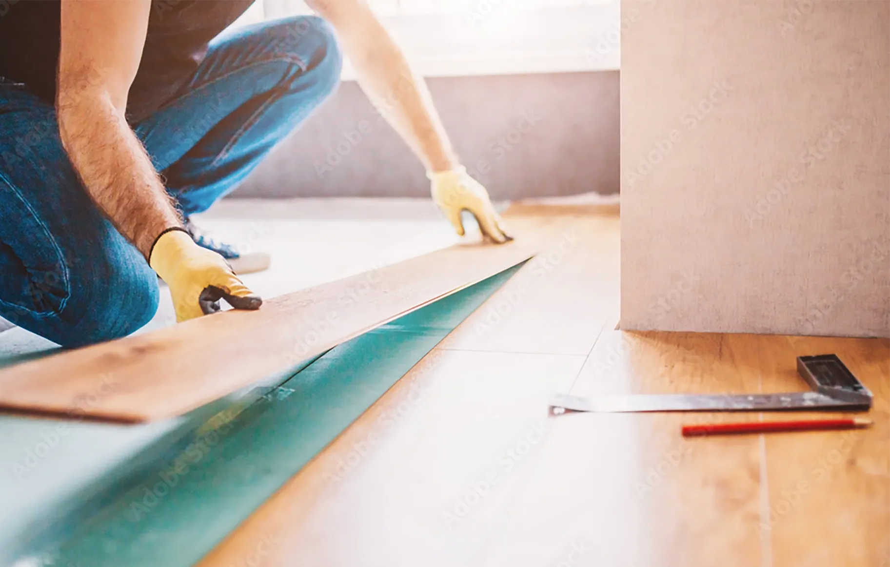 A person wearing yellow gloves installs wood laminate flooring, with a metal square and pencil lying on the floor nearby.
