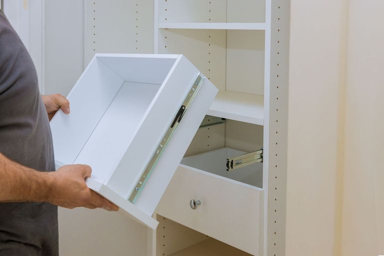 A person in a gray shirt installs a white drawer into a cabinet unit, aligning the metal drawer slides.