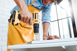 A person wearing a yellow apron uses a cordless drill to attach a metal hinge to a white furniture panel.