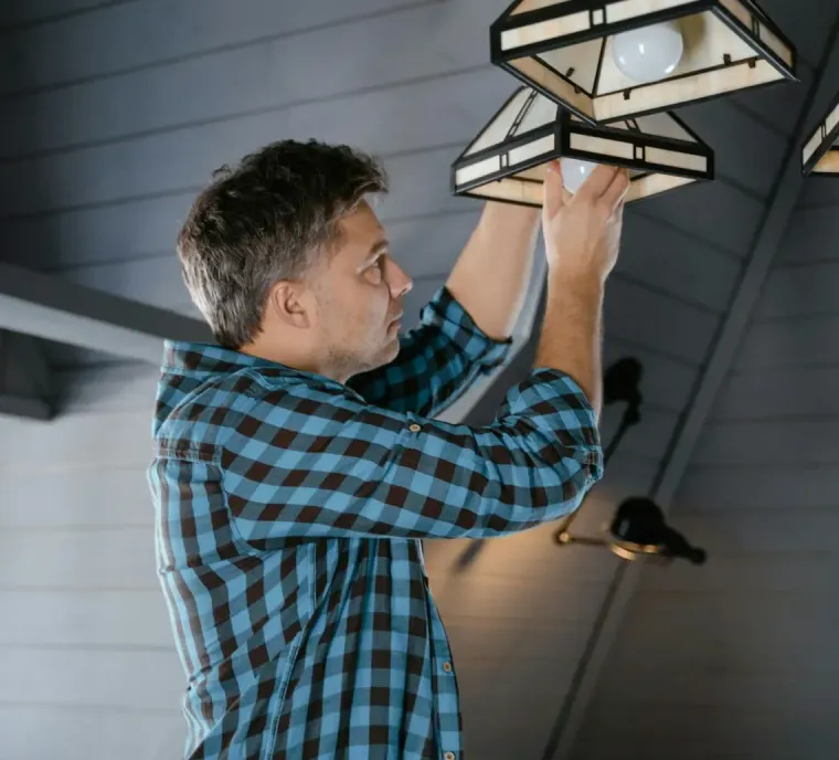 A person in a blue and black plaid shirt stands on a ladder, reaching up to change a lightbulb in a hanging lamp.