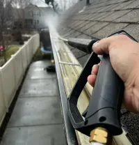 A first-person view of a hand using a pressure washer to clean a dirty residential gutter along a roofline.