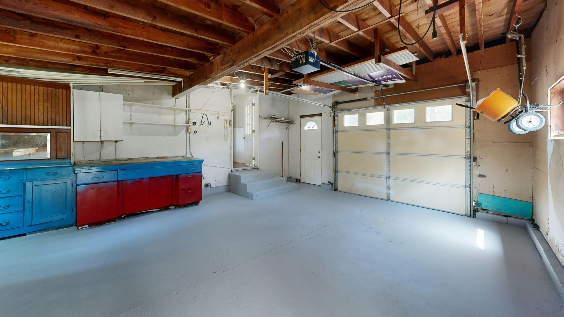 An empty garage with concrete floors, wooden ceiling beams, blue and red cabinets, and a white garage door.