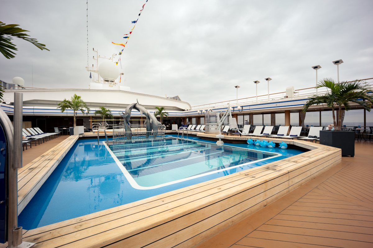 Pool deck on a cruise ship with a rectangular pool, lounge chairs, and palm trees under a cloudy sky.