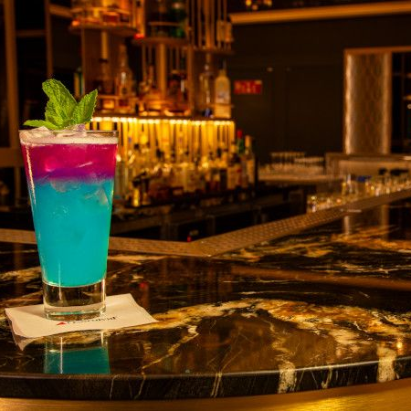 Cocktail with blue and purple layers garnished with mint on a bar. The bar is dark with a gold marble top.
