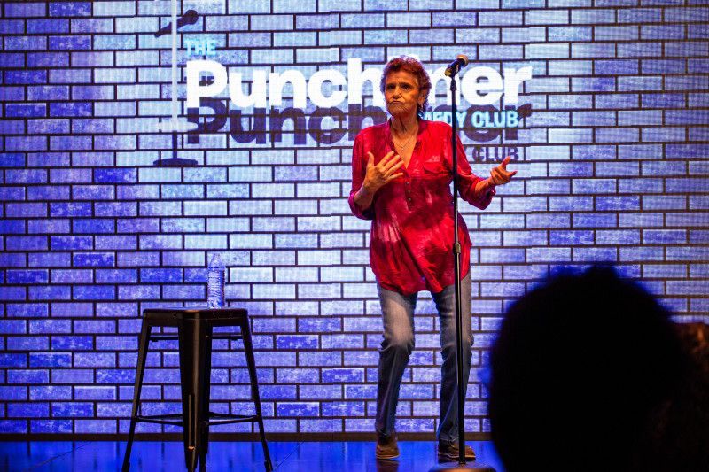 Woman on stage at comedy club, red top, jeans, gestures with hands, brick backdrop, logo.