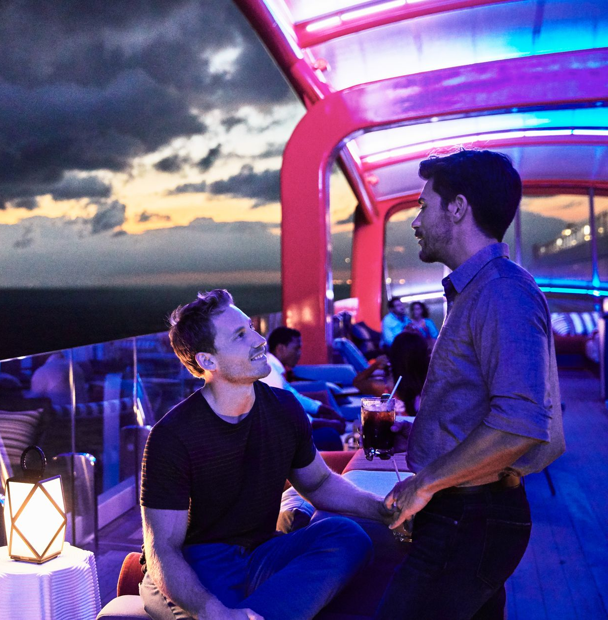 Two men conversing on a deck overlooking water at sunset. Dim lighting and dark clouds.
