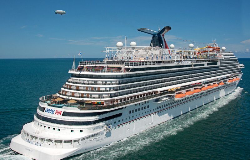 Cruise ship sailing on a blue sea under a clear sky; a blimp flies nearby.