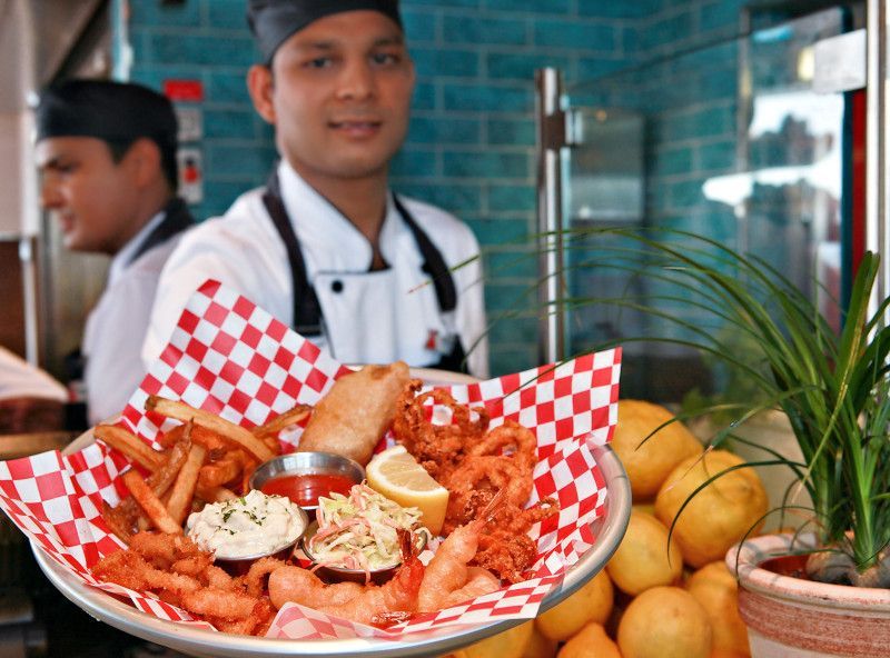 A restaurant worker holds a platter of fried food, smiling. Another worker is in the background.