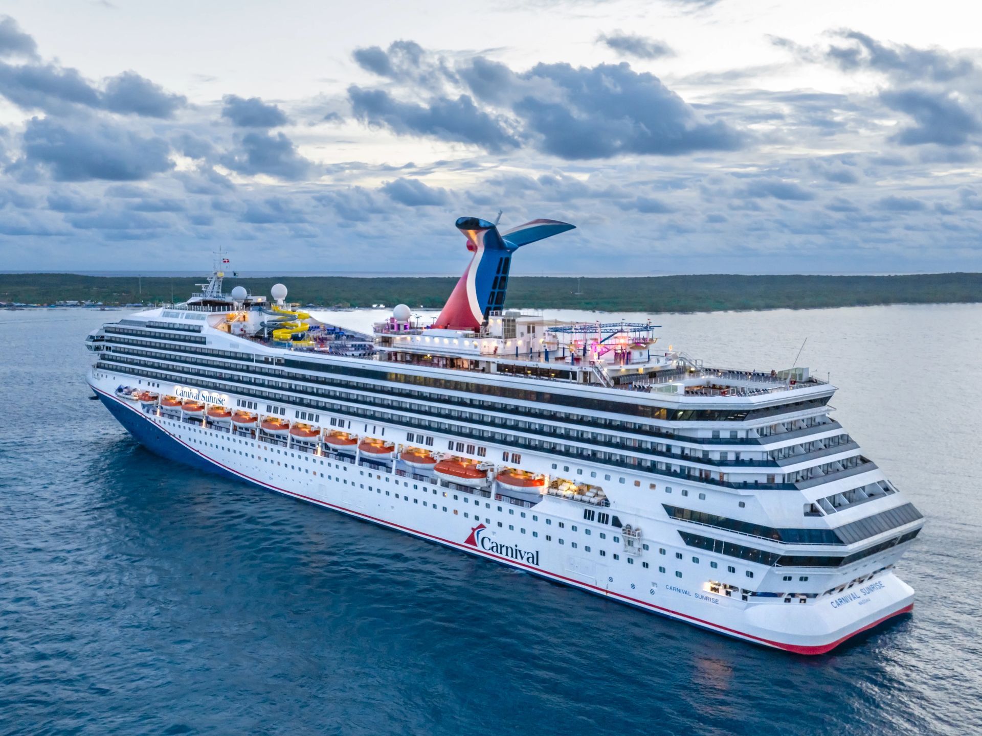 Large Carnival cruise ship on the ocean, blue and white hull, red and blue funnel, cloudy sky.