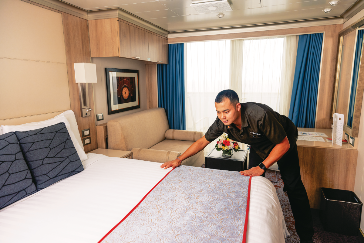 Man making a bed in a cruise ship cabin. He is arranging a decorative bed runner.