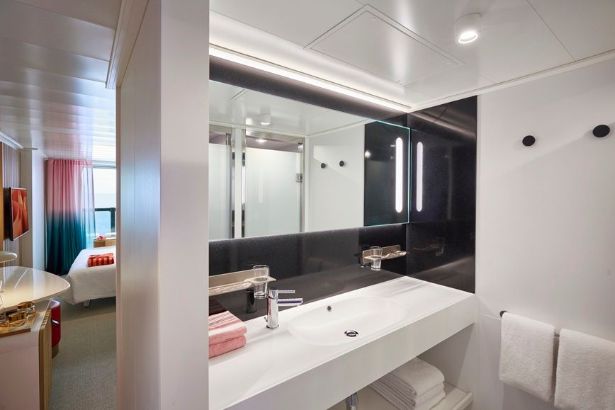 Modern bathroom with large mirror, sink, towels, and a glimpse of a nearby bedroom.