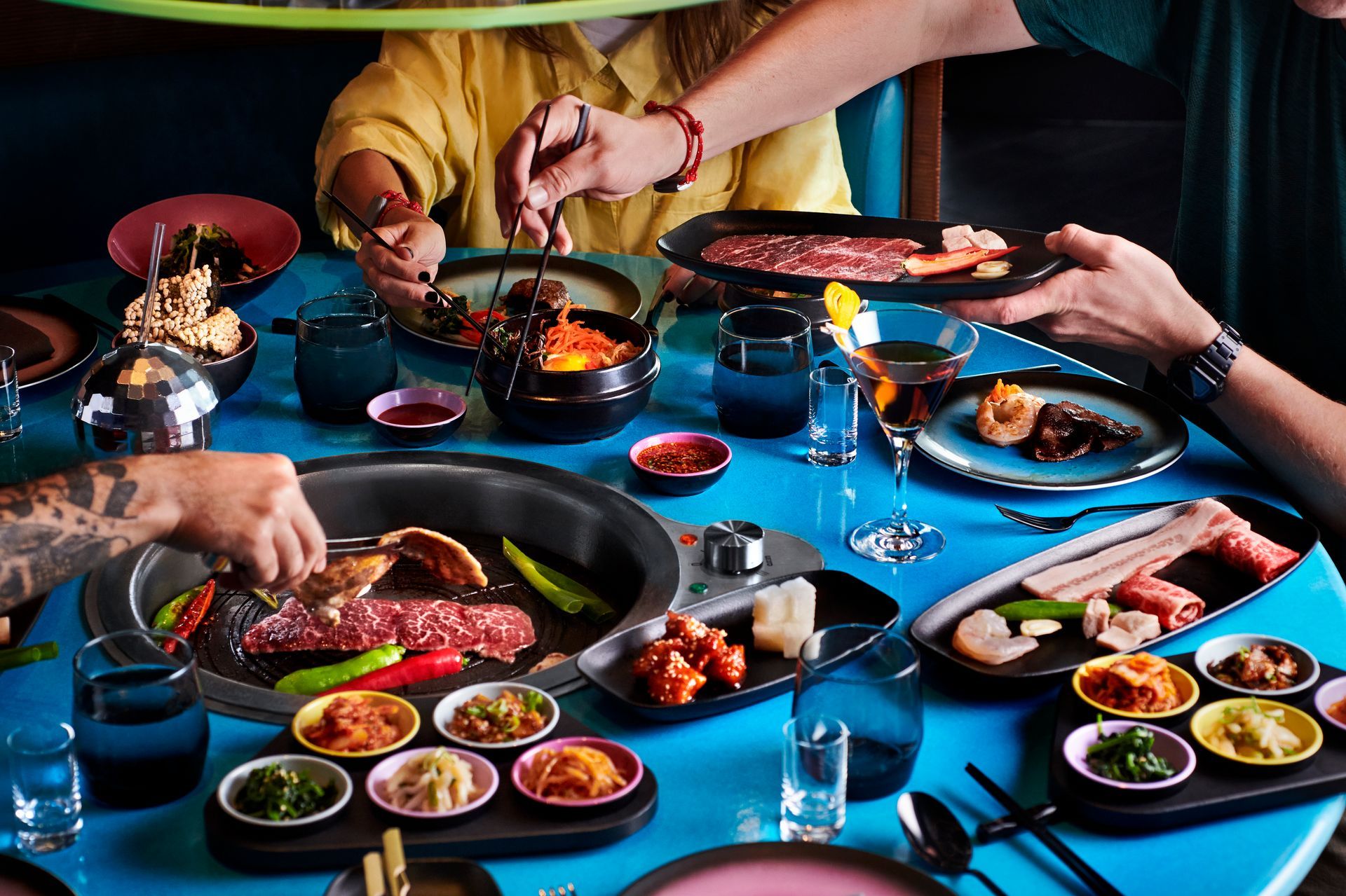 People enjoying Korean BBQ at a blue table. Raw meat, side dishes, and a grill are present.