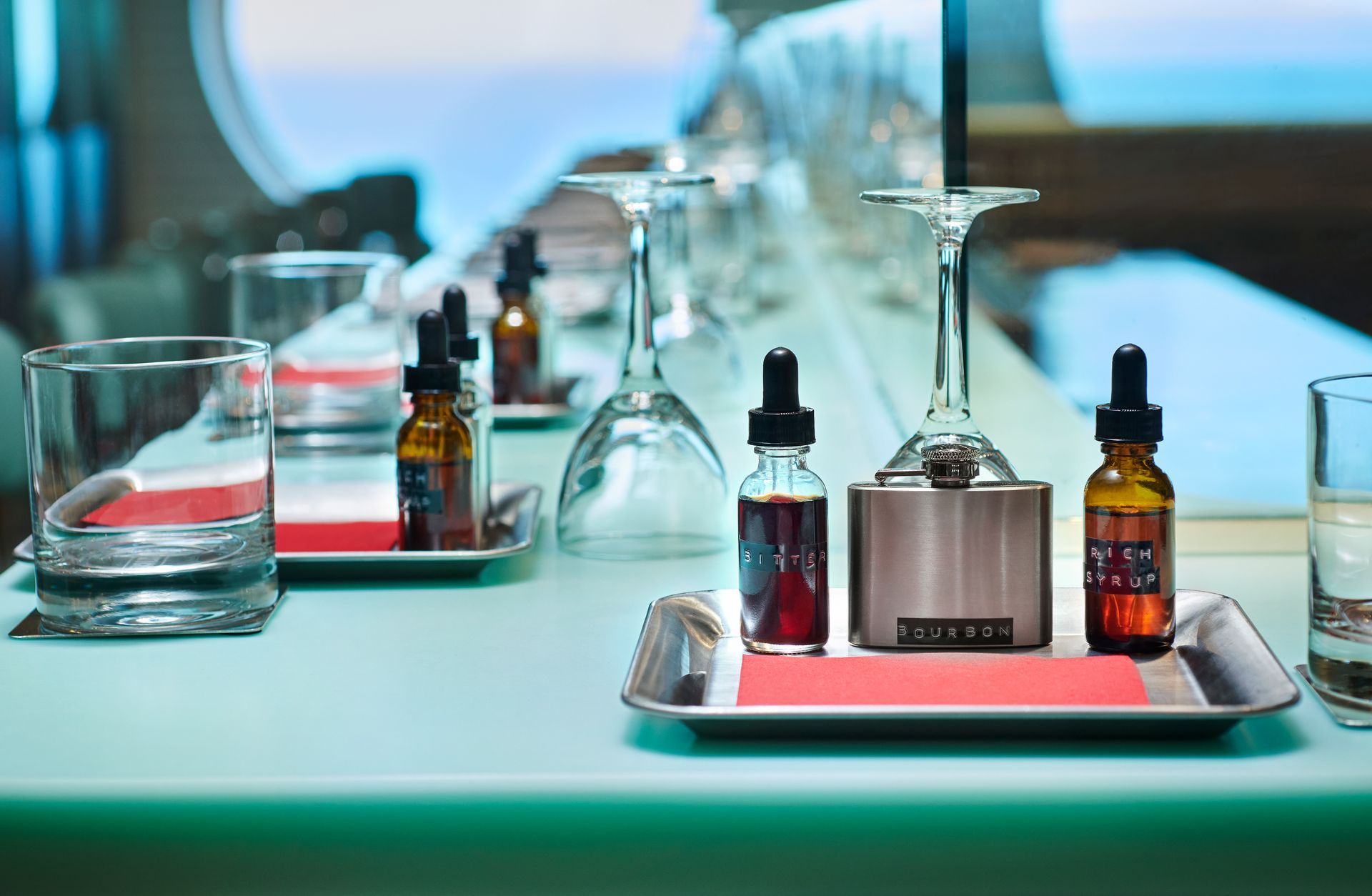 Bottles with droppers, small metal device, and glasses on a counter, possibly for mixing drinks, near a window.