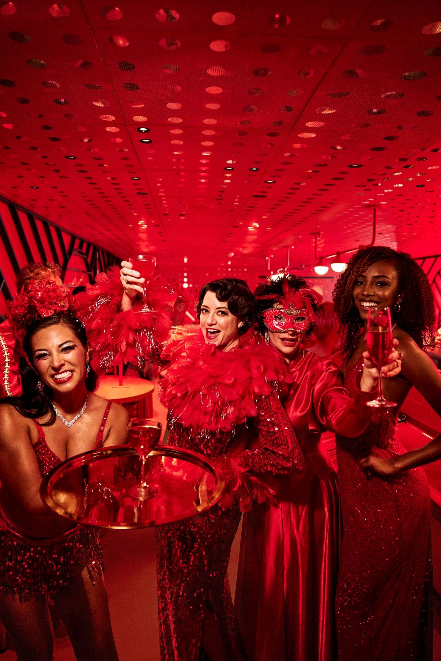 Four people in red attire at a party, holding drinks and smiling under a red-lit ceiling.