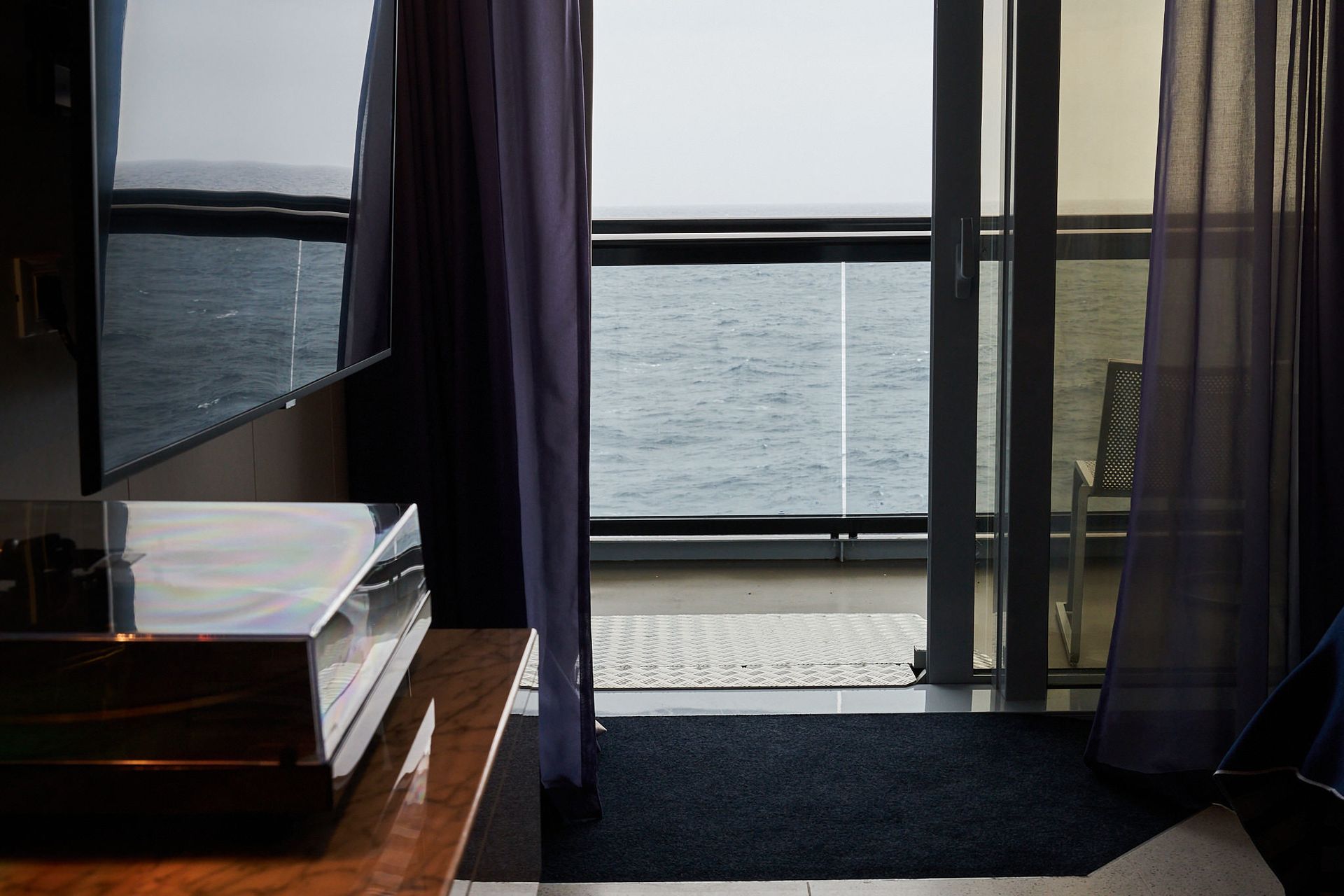 Balcony view: dark curtains frame ocean scene; TV, dresser in foreground.