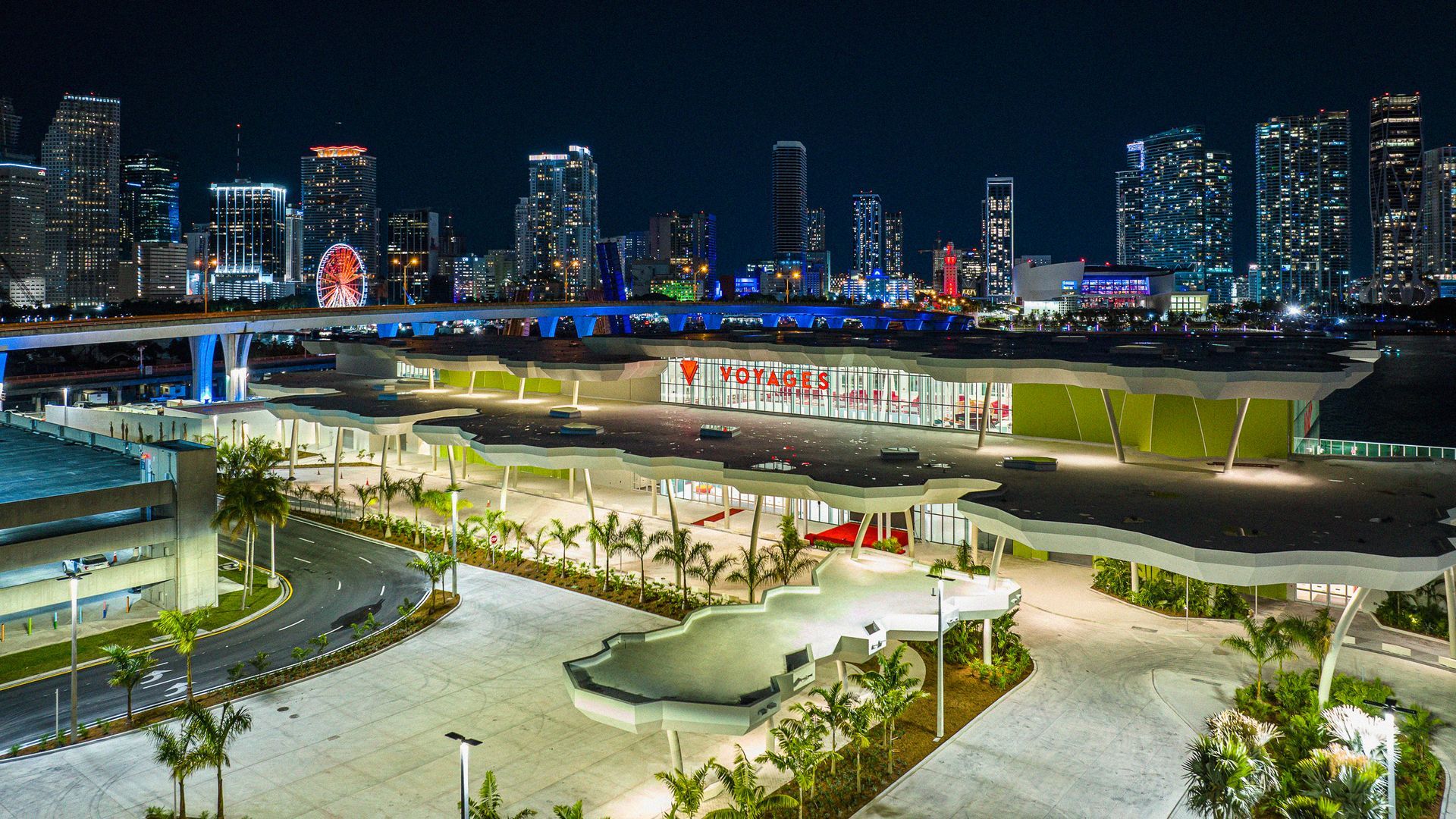 Nighttime cityscape of Miami with illuminated buildings, waterfront, and palm trees.