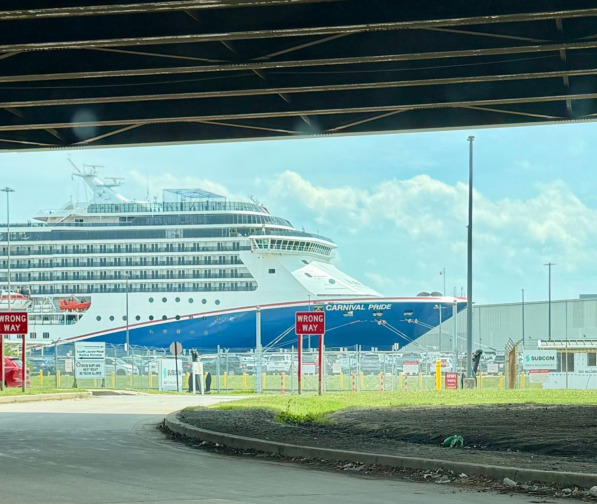 Large white cruise ship docked at a port beneath an overpass, seen across a fenced roadway.