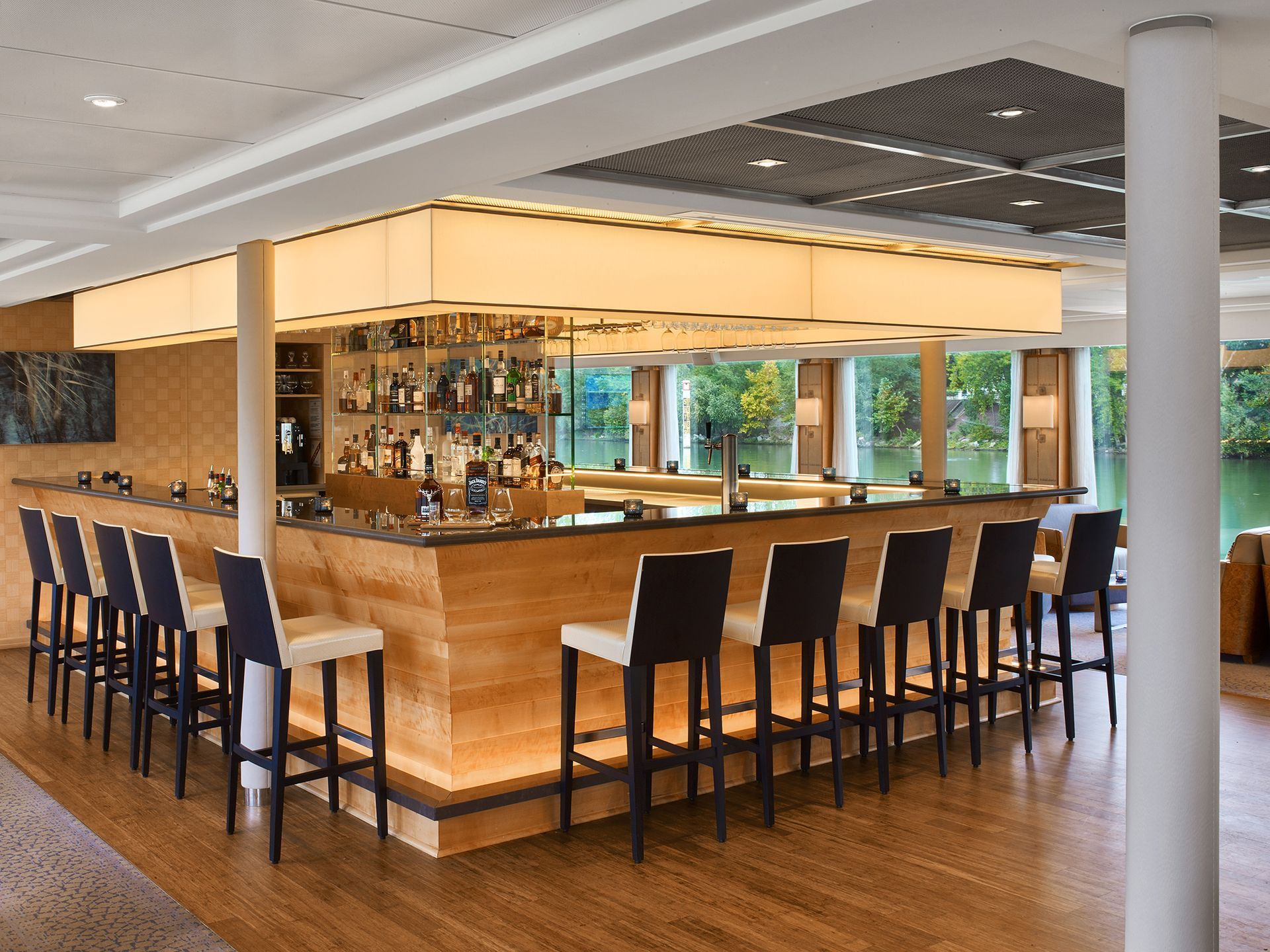 Bar with wooden facade and stools, drinks, and bright lighting, with river view.