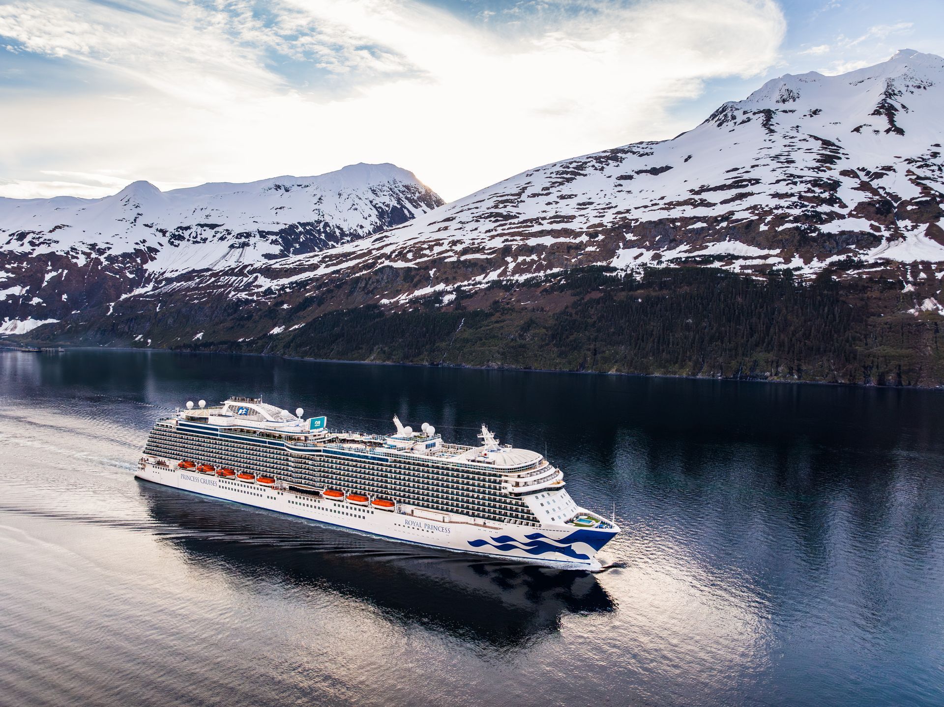 A Princess Cruises ship sails through calm, reflective waters surrounded by towering snow-capped mountains in Alaska. The large white vessel, with blue wave graphics on its hull, glides beneath a bright sky with soft clouds, showcasing the serene beauty of Alaskan wilderness and glacier-fed fjords.