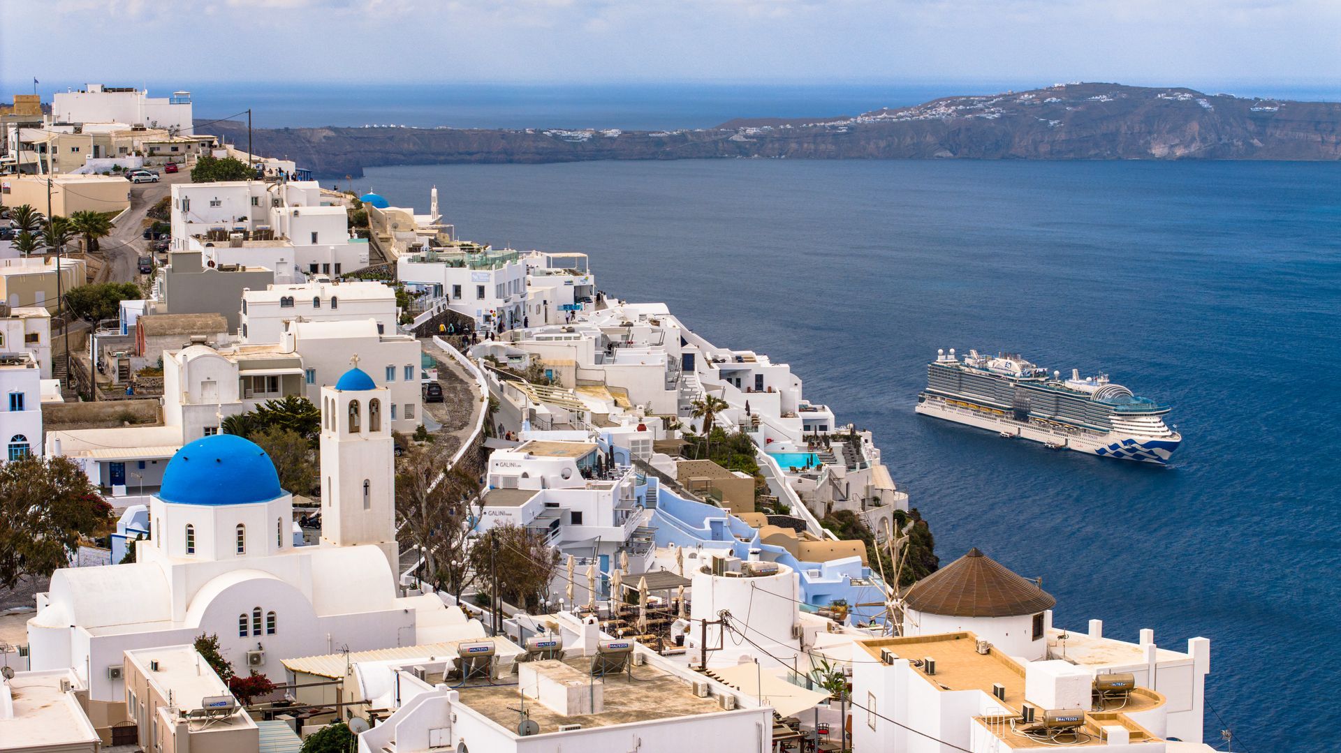 Fira, Santorini cliffside village with blue-domed buildings overlooks the Aegean Sea and the Sun Princess cruise ship.