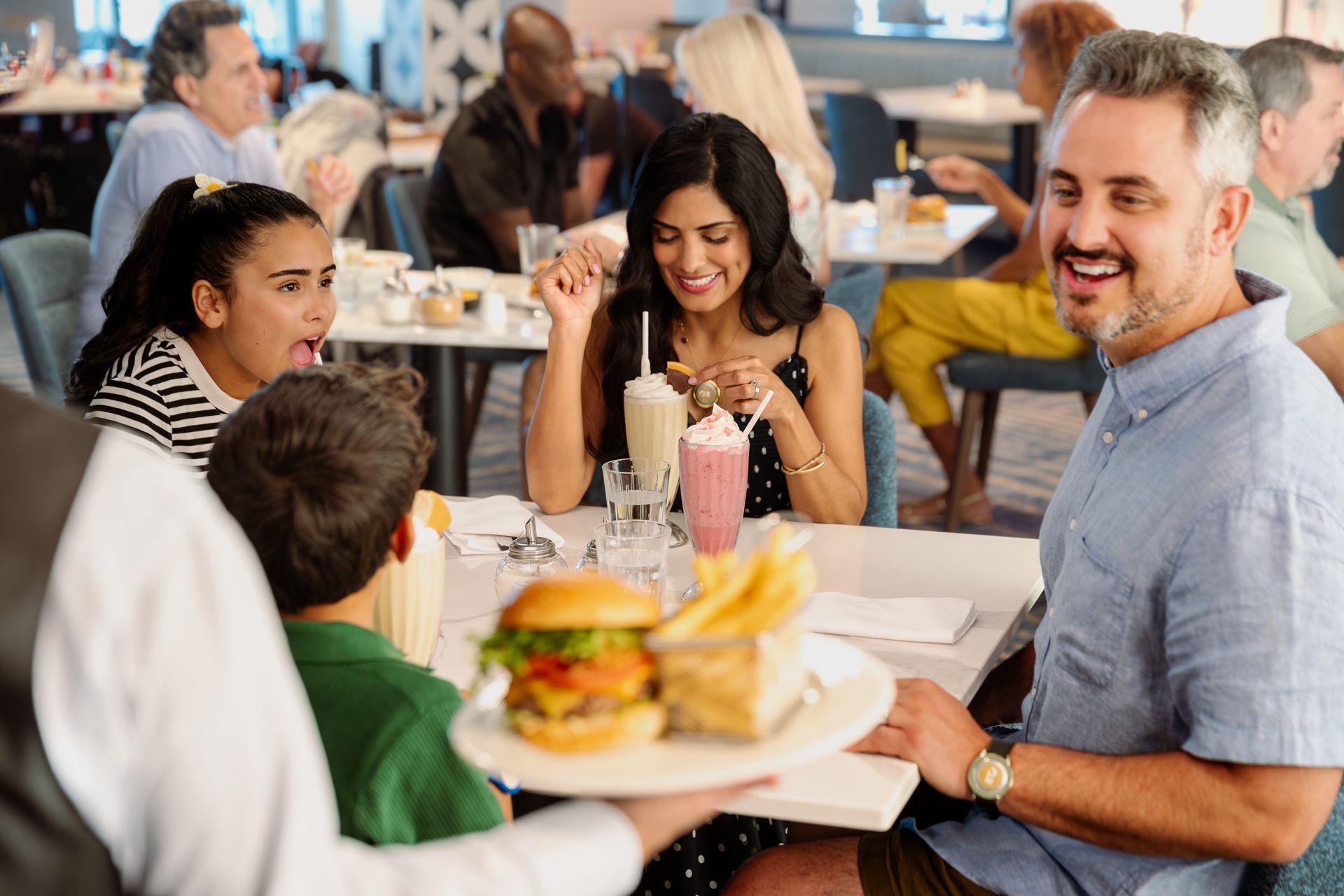  A joyful family is seated at a table in a bright restaurant. A waiter delivers a burger and fries as the father smiles, and the daughter reacts excitedly.