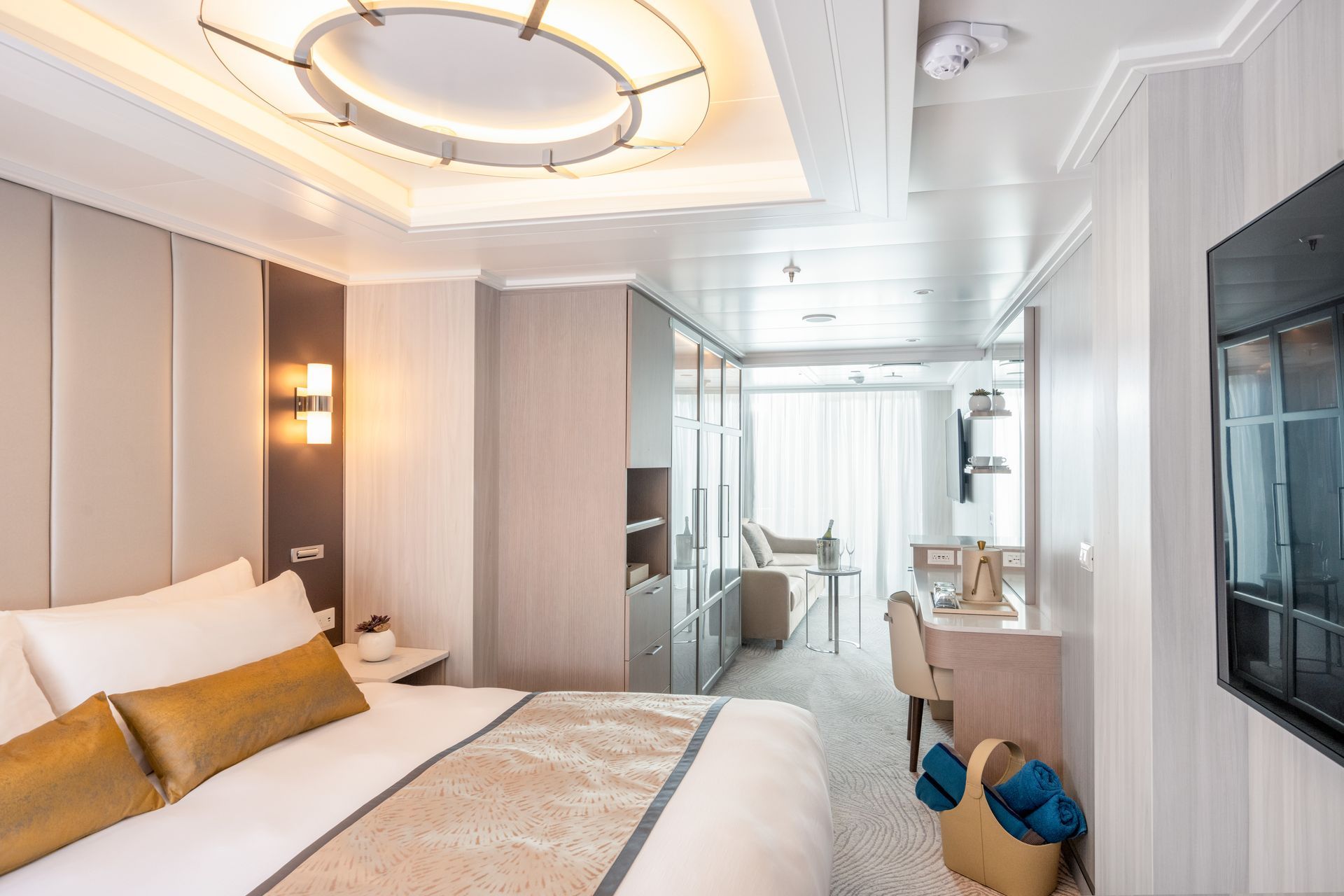 Luxury cruise ship stateroom, neutral color palette, bed, desk, seating area, large window, and recessed ceiling lighting.