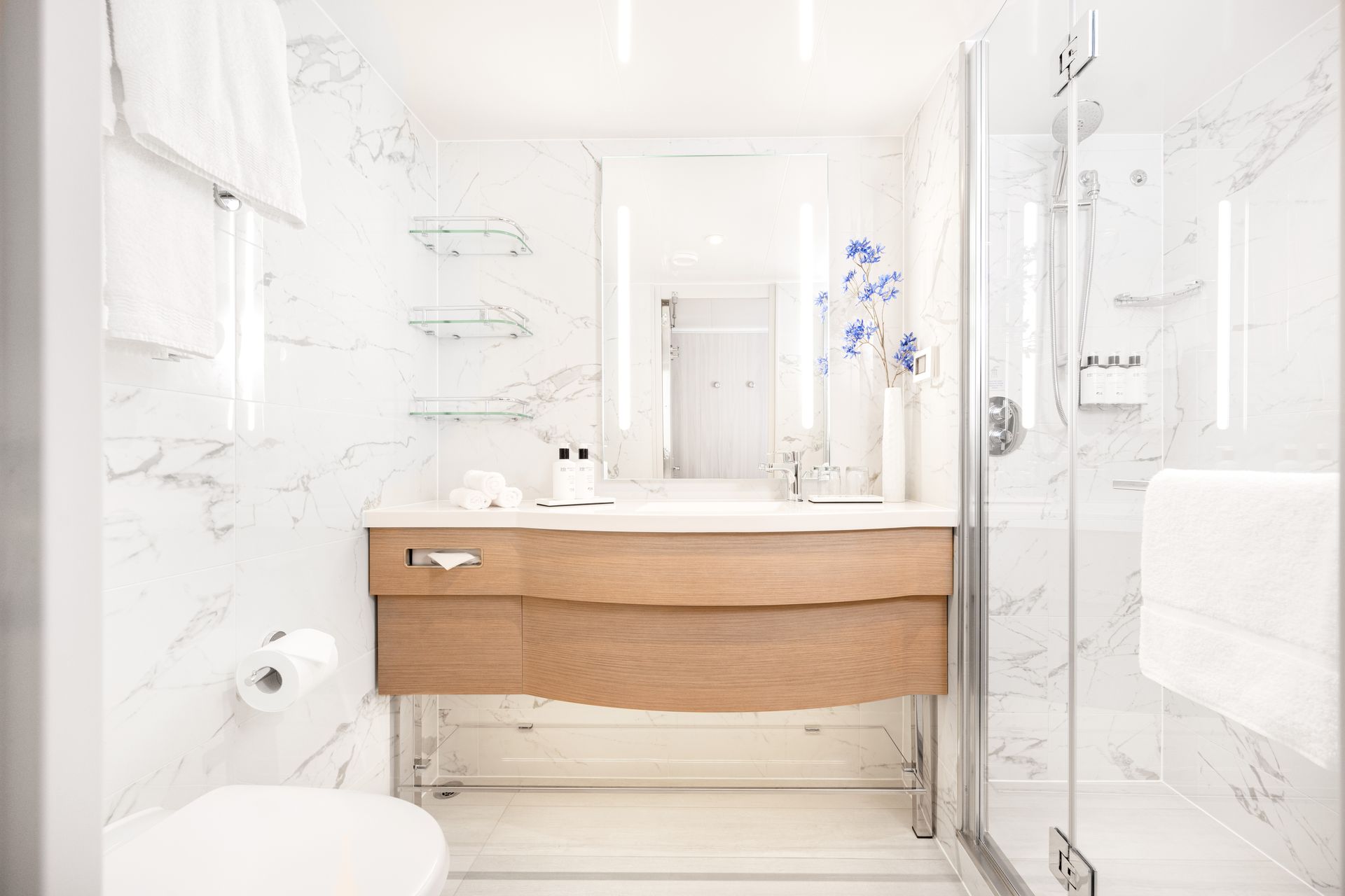 Bright, modern bathroom with marble walls, a wooden vanity, a glass shower, and white towels.