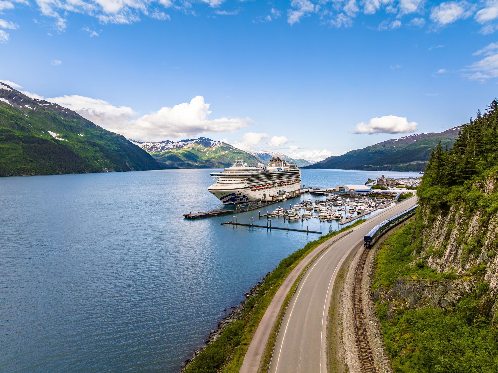Cruise ship docked in a harbor, train running along a coastal road, mountains in background, blue sky.