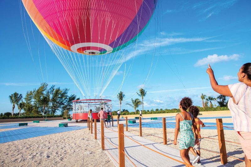 People watching a colorful hot air balloon launch from a beach area with a trolley in the background.